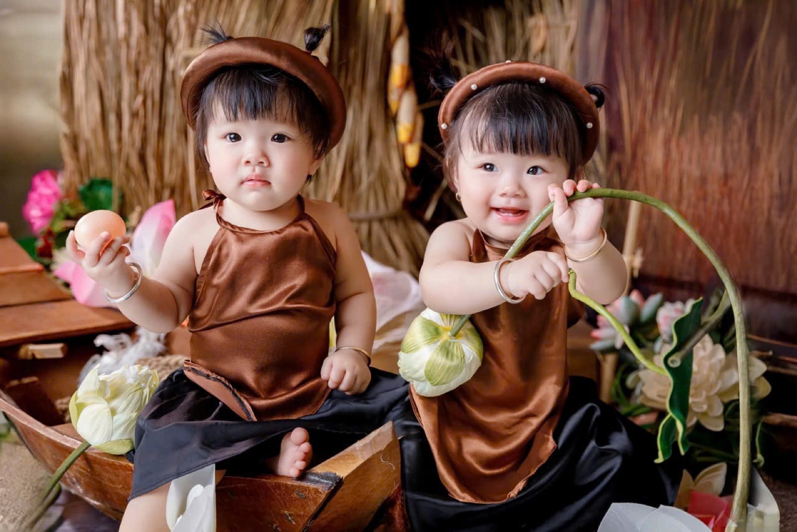 Gợi ý chụp ảnh Tết cho bé: hai bé gái mặc yếm nâu, váy đen, đội mấn nâu, ngồi trên xuồng gỗ giữa bông sen, một bé cầm quả trứng, một bé cầm cành sen cười tươi, phía sau là vách rơm