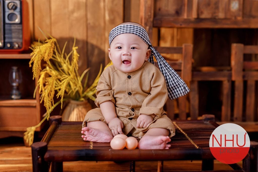 Chụp ảnh bé 6 tháng ngồi vững trên phản tre, cười hở lợi nhìn máy