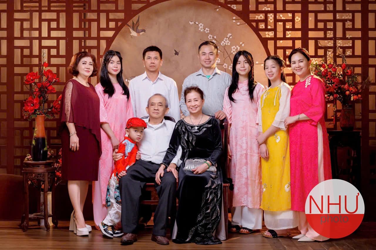 Ảnh gia đình áo dài đông người trên nền vách gỗ: bà mặc áo dài nhung đen và ông mặc sơ mi trắng ngồi ghế, bé trai áo đỏ đội mũ đỏ đứng tựa vào ông, các cô gái đứng sau mặc áo dài hồng, vàng và hồng sen