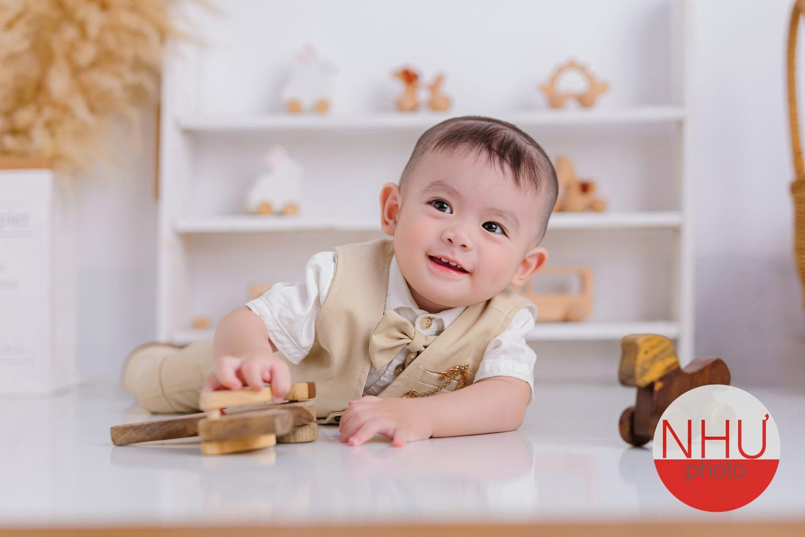 In ảnh cho bé: em bé trai mặc sơ mi trắng, gi-lê màu be thắt nơ, nằm sấp trên sàn trắng cười nhìn lên, tay cầm đồ chơi gỗ, phía sau là kệ trắng bày đồ chơi gỗ