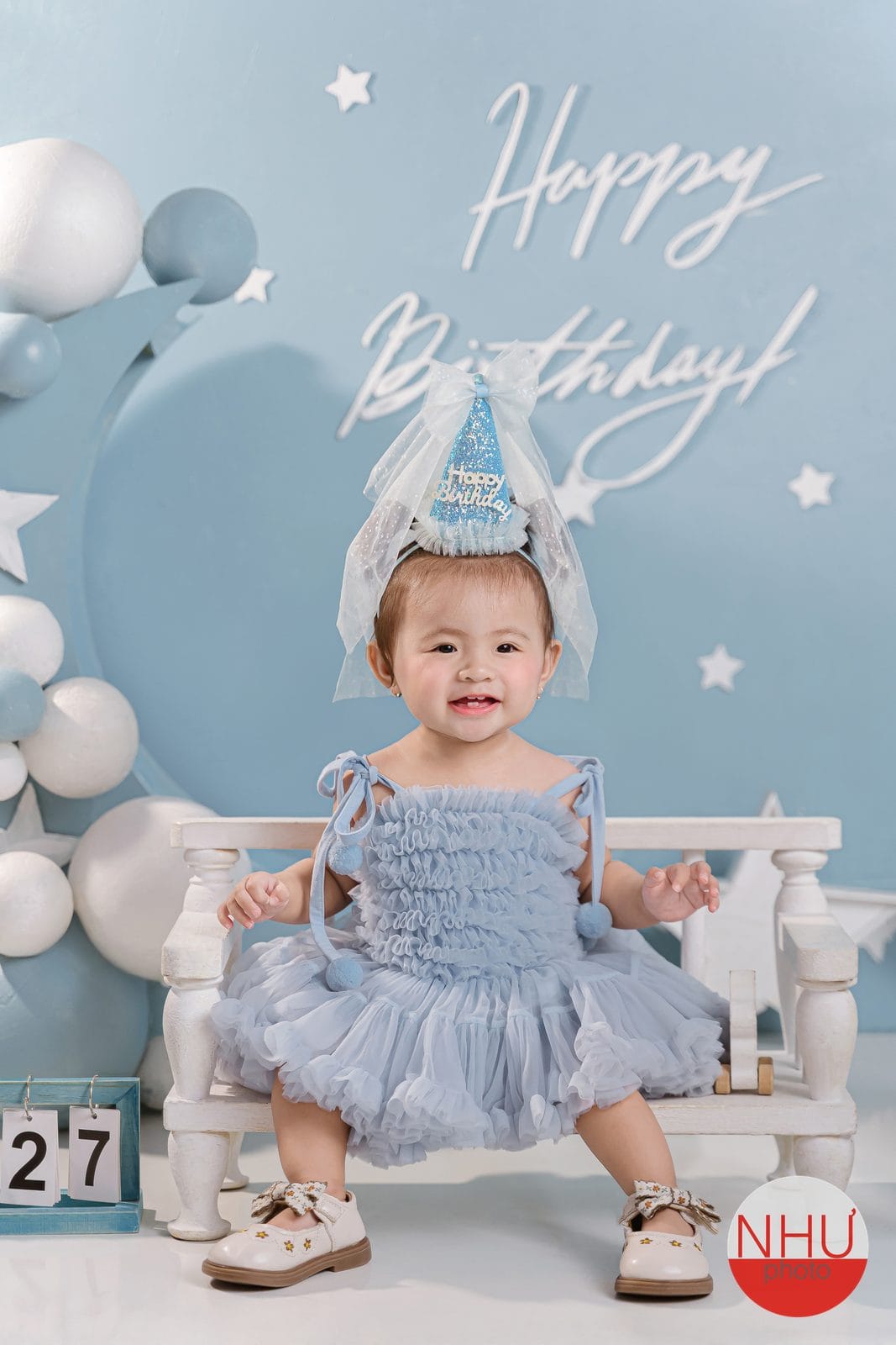 Studio chụp ảnh cho bé: bé gái đầm xòe xanh, đội nón sinh nhật có voan, ngồi trên ghế băng trắng, phông xanh có chữ Happy Birthday