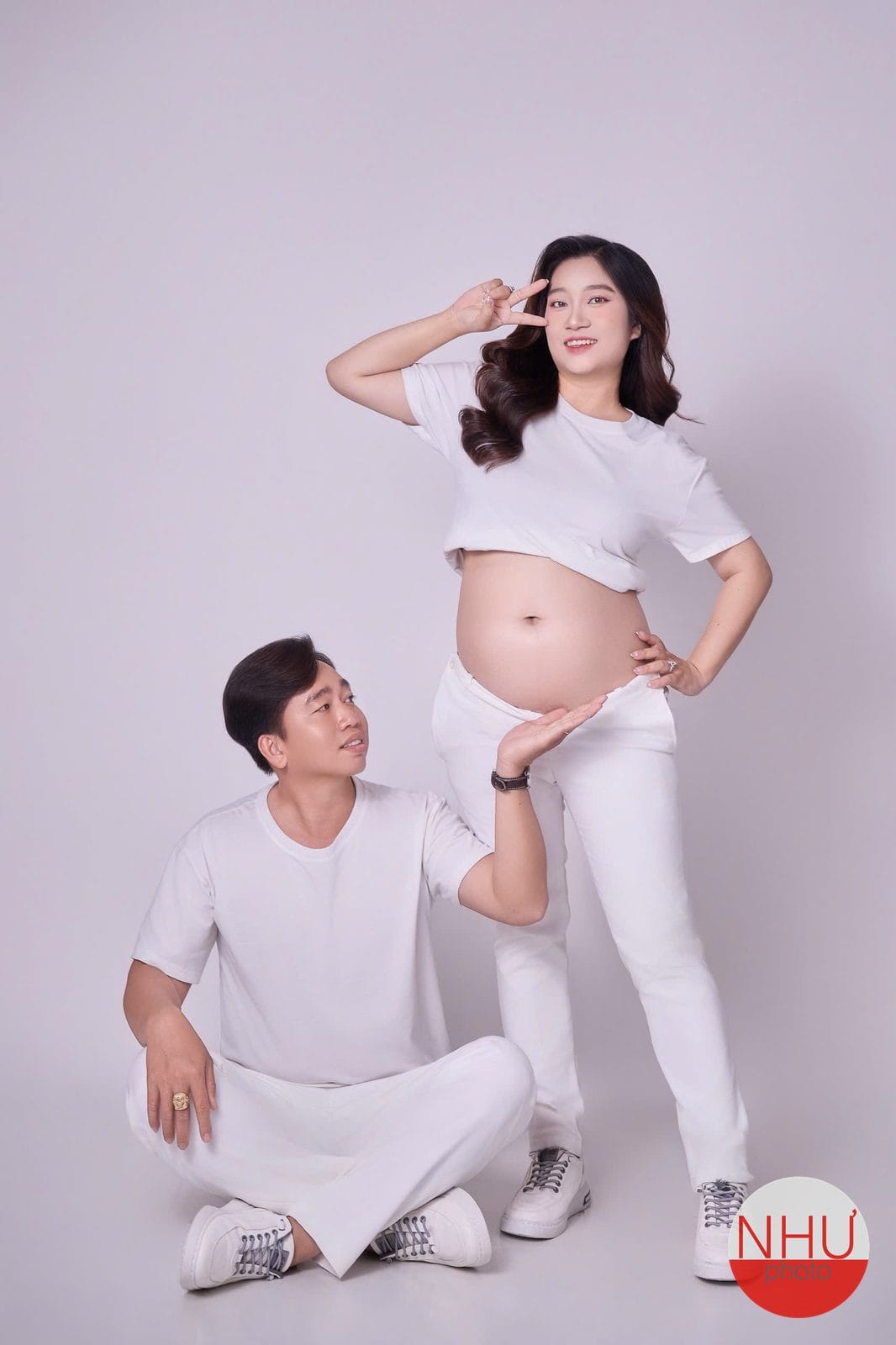 Chụp ảnh bầu tại studio: mẹ bầu mặc áo thun trắng ngắn để lộ bụng, một tay giơ chữ V cạnh mặt, một người đàn ông mặc đồ trắng ngồi dưới sàn đưa tay hướng về bụng mẹ