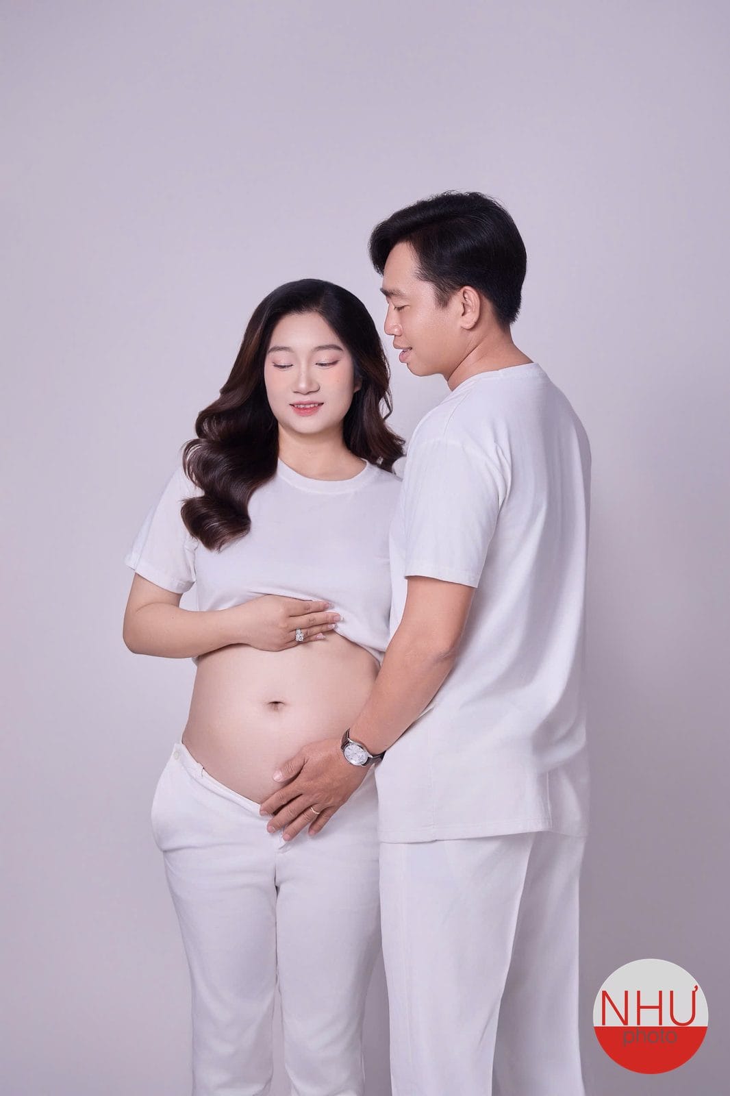 Chụp ảnh bầu tại studio: mẹ bầu mặc đồ trắng nhắm mắt, một tay vén áo đặt lên bụng, một người đàn ông mặc đồ trắng đặt tay lên bụng mẹ