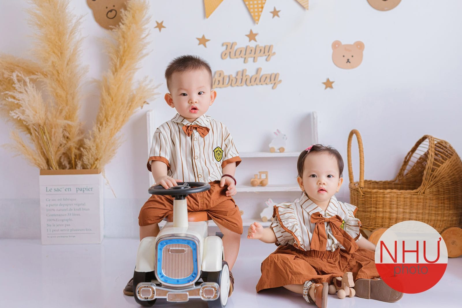 Chụp ảnh bé: bé trai ngồi trên xe chòi chân màu trắng, bé gái mặc đồ nâu ngồi bệt bên cạnh, phía sau có chữ Happy Birthday