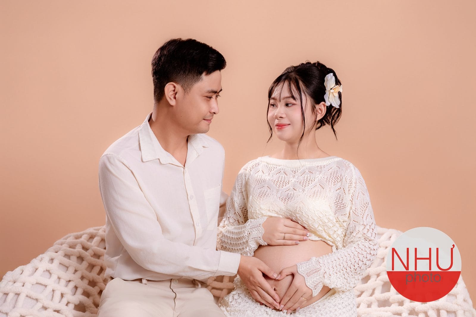 Chụp ảnh bầu cute cùng chồng: hai vợ chồng mặc đồ trắng kem ngồi trên chăn len đan to, bàn tay hai người ghép thành hình trái tim trên bụng bầu, quay sang nhìn nhau