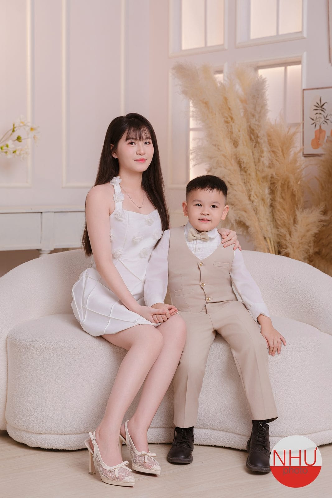 Chụp ảnh mẹ và bé trai: mẹ mặc váy trắng ngồi trên ghế sofa trắng, con trai khoảng năm tuổi mặc gile màu be thắt nơ ngồi sát bên, phía sau là bông lau