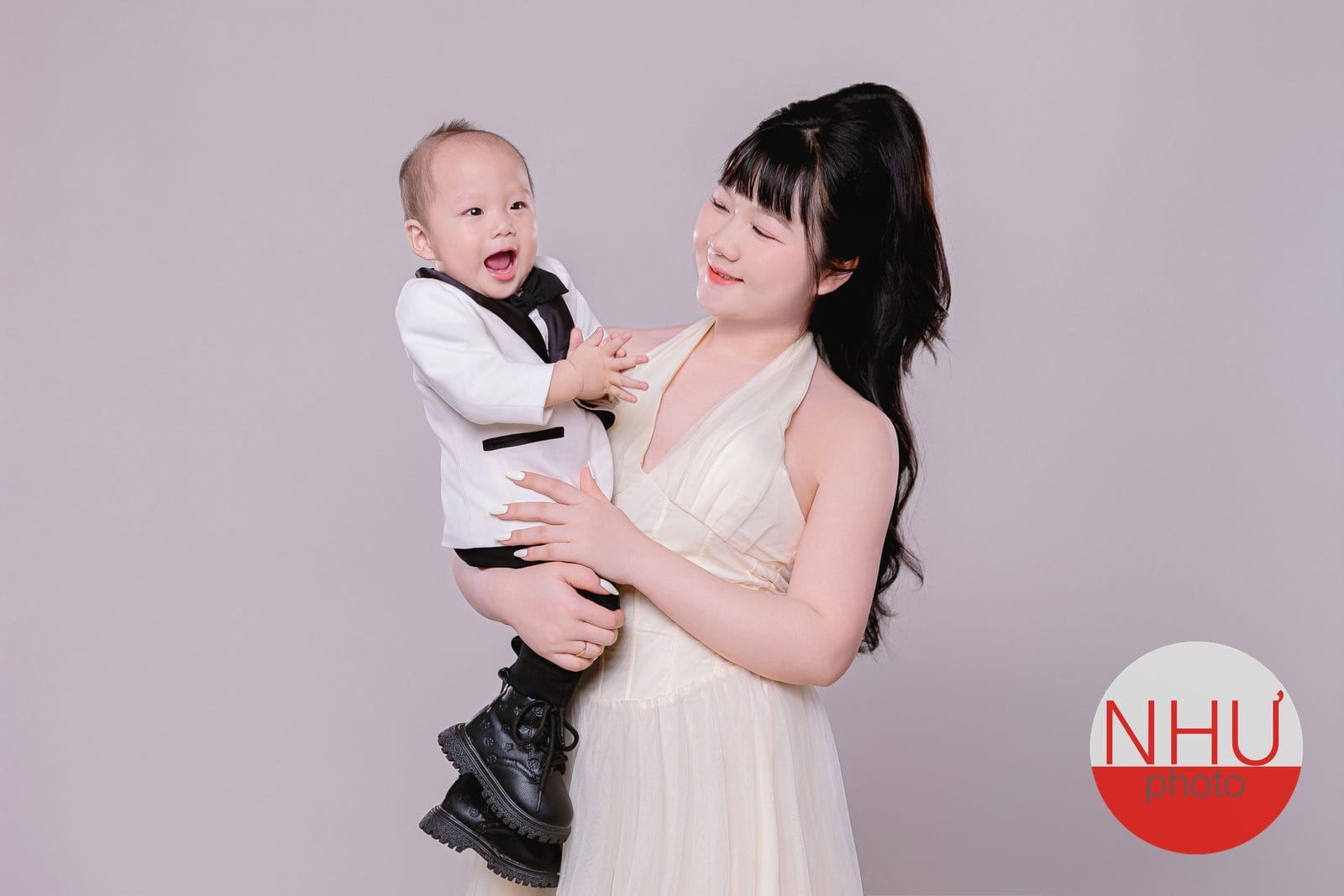 Chụp ảnh mẹ và bé trai: mẹ mặc váy dài màu kem, tóc buộc cao, bế con trai nhỏ mặc vest trắng nơ đen, con cười toe, mẹ nghiêng đầu nhìn con
