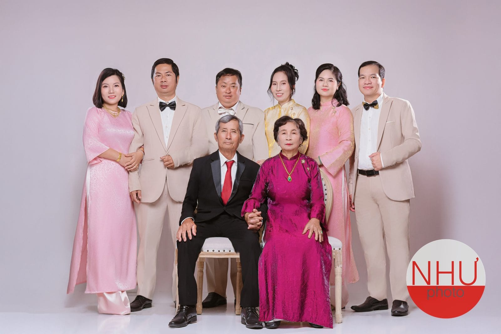 Chụp ảnh gia đình áo dài: ông mặc vest đen cà vạt đỏ và bà mặc áo dài tím sen ngồi ghế nắm tay nhau, sáu người đứng phía sau, ba người phụ nữ mặc áo dài hồng và vàng, ba người đàn ông mặc vest be