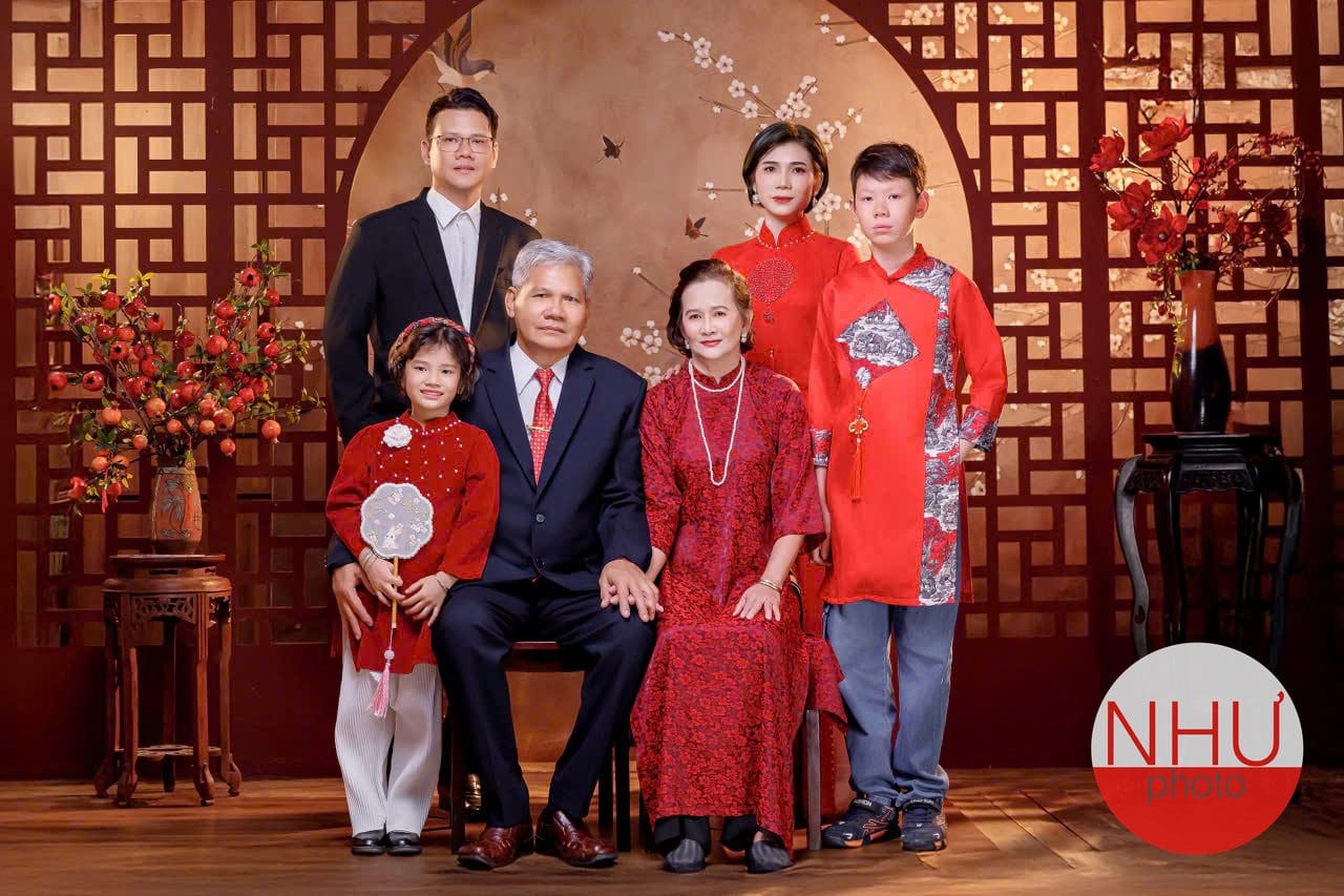 Chụp ảnh gia đình áo dài trên nền vách gỗ đỏ có lọ hoa đỏ: ông vest xanh đen và bà áo dài ren đỏ ngồi ghế phía trước, bé gái áo đỏ cầm quạt tròn, người phụ nữ áo dài đỏ, cậu cháu mặc áo dài cách tân đỏ với quần jean và giày thể thao