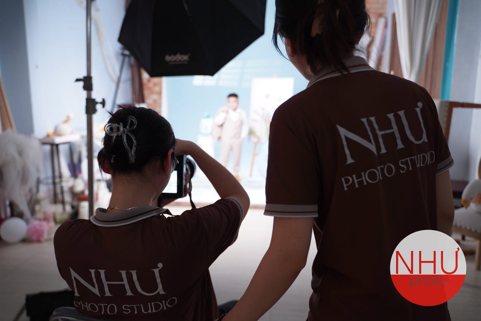 Hậu trường studio: hai thợ mặc áo nâu in chữ NHƯ Photo Studio đứng quay lưng, một người cầm máy chụp người đàn ông mặc vest trong set phía trước