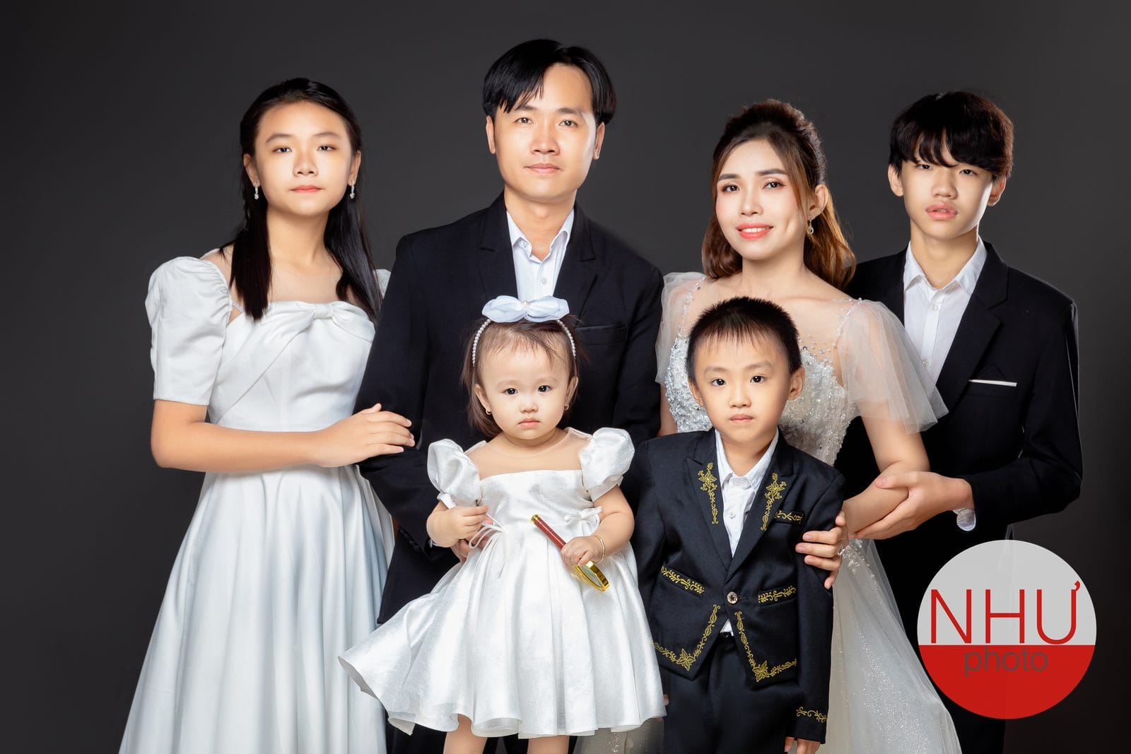 Ảnh gia đình sáu người chụp studio trên nền xám đậm: hai người lớn, hai thiếu niên và hai bé nhỏ mặc đồ trắng đen đứng sát nhau