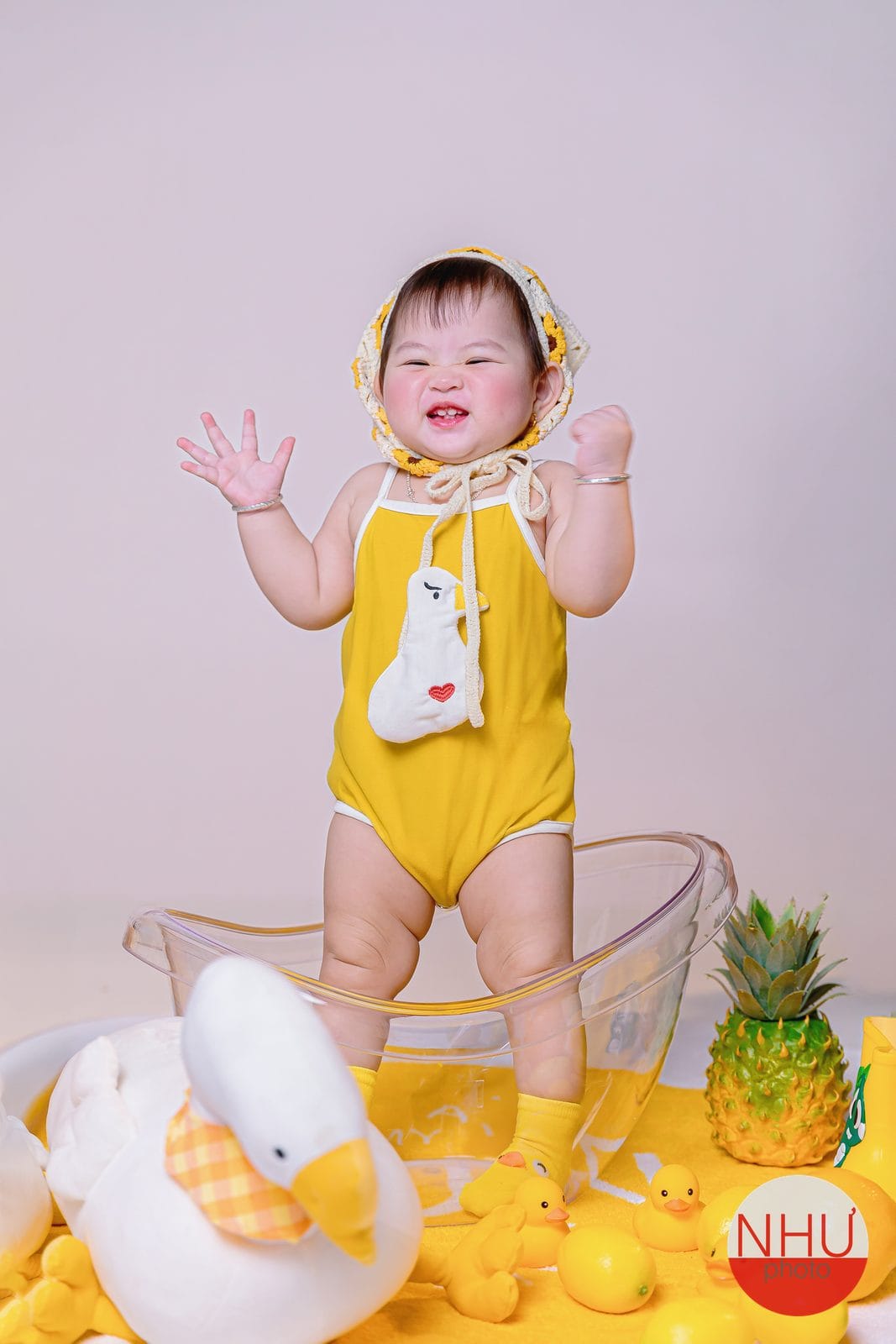 In ảnh photobook cho bé: em bé mặc đồ liền thân vàng có hình ngỗng trắng, đội khăn len đan hoa hướng dương, đứng giơ hai tay cười trong chiếc chậu nhựa trong, quanh chân là thú bông ngỗng trắng, vịt cao su vàng và trái thơm
