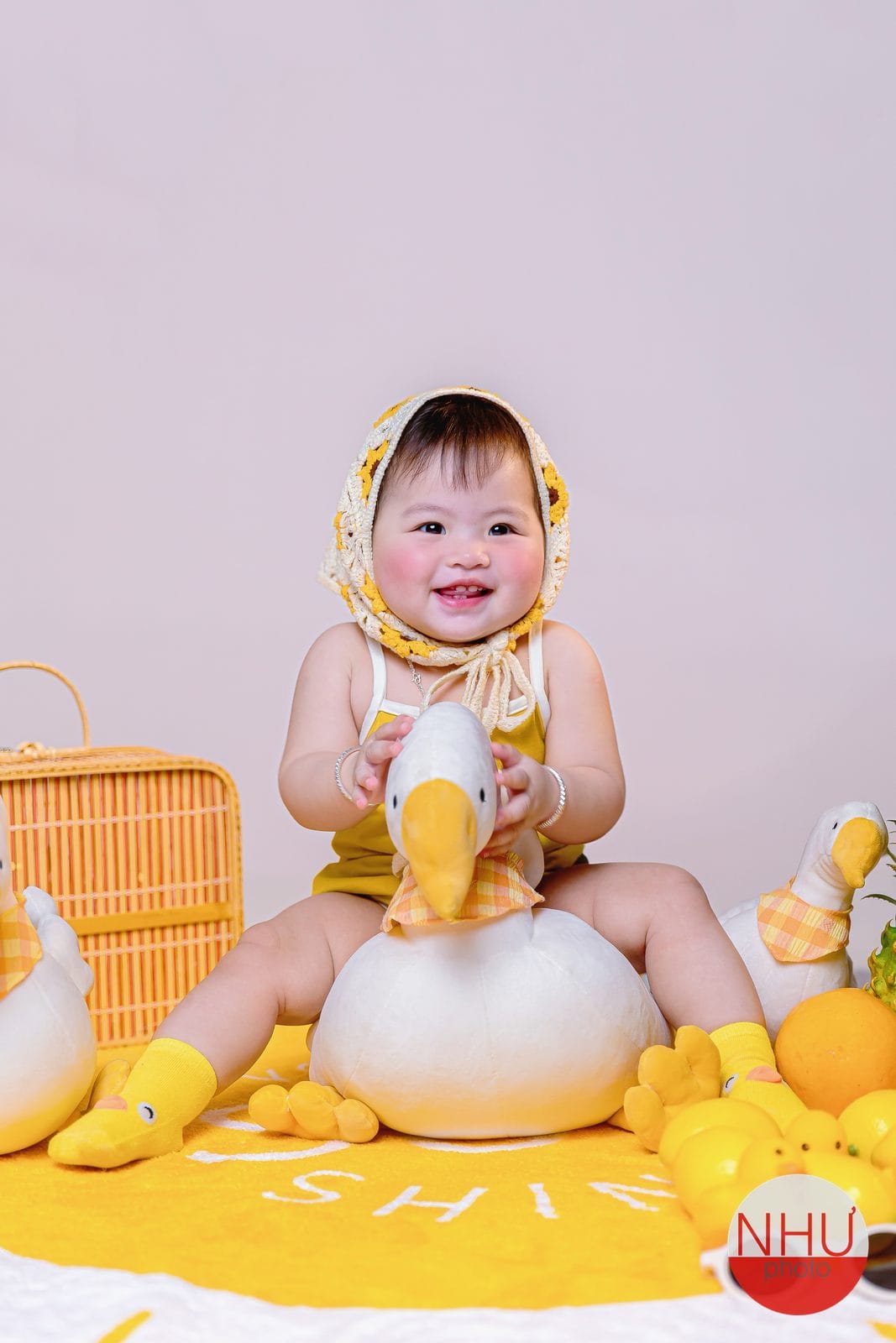In ảnh photobook cho bé: em bé đội khăn len đan hoa hướng dương, mặc đồ vàng, ngồi trên thảm vàng ôm cổ một con ngỗng bông trắng lớn, cười tươi, bên cạnh là giỏ mây và trái cây màu vàng