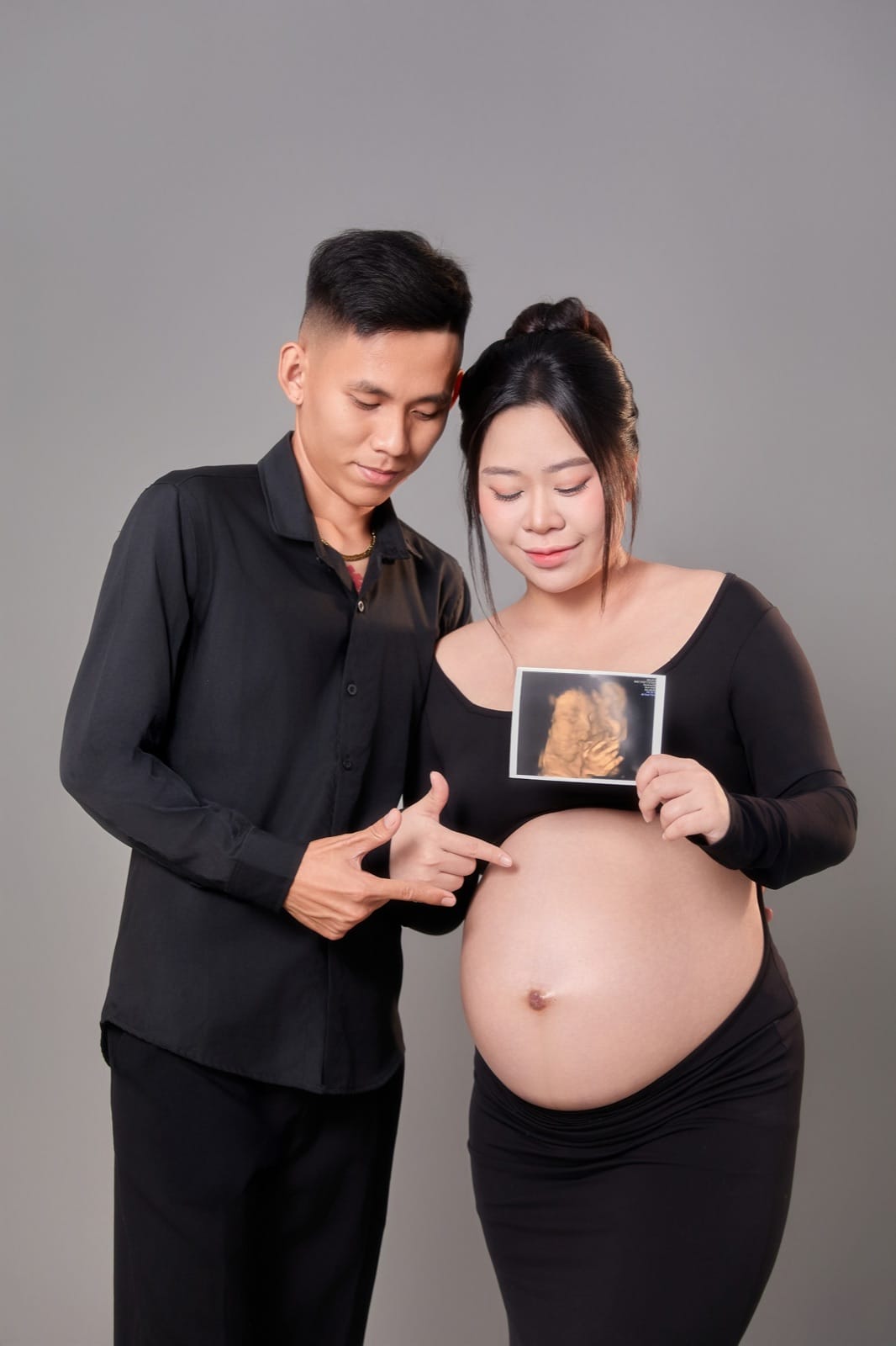 Chụp ảnh bầu cute cùng chồng trên phông xám: hai vợ chồng mặc đồ đen cùng nhìn xuống bụng, chồng dùng hai tay chỉ vào bụng mẹ, mẹ cầm ảnh siêu âm trước ngực