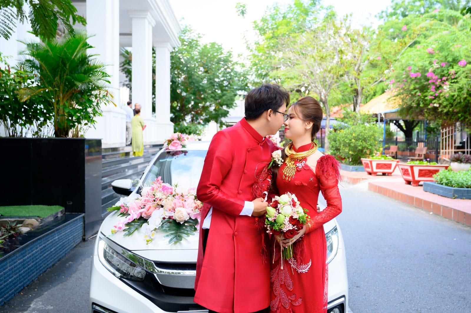 In ảnh cưới: cô dâu chú rể mặc áo dài đỏ đứng trước xe hoa trắng kết hoa hồng, cô dâu cầm bó hoa sen và hồng đỏ, phía sau là hàng cây xanh và bậc thềm có khách