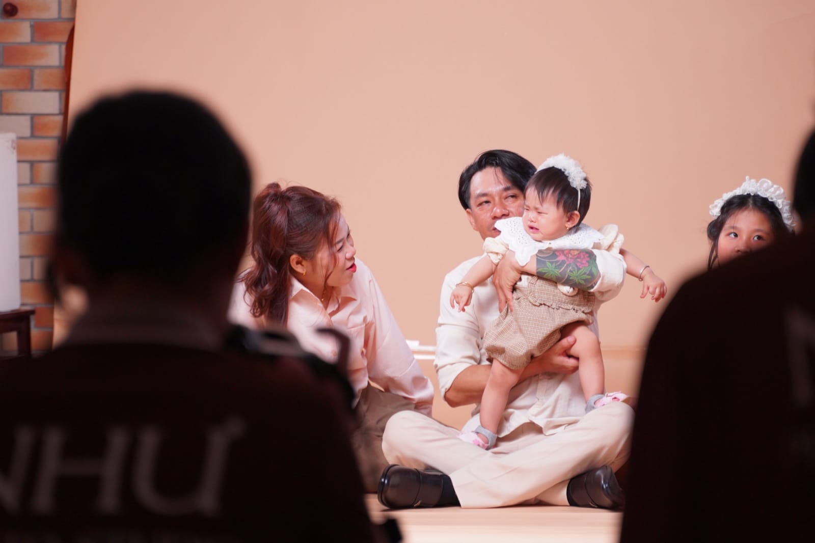 Hậu trường buổi chụp ảnh cho bé và gia đình: người đàn ông ngồi xếp bằng bế bé gái đội băng đô ren trắng đang mếu, người phụ nữ ngồi cạnh nhìn bé, một bé gái lớn hơn ở mép phải, lưng ê-kíp mờ phía trước có áo in chữ NHƯ