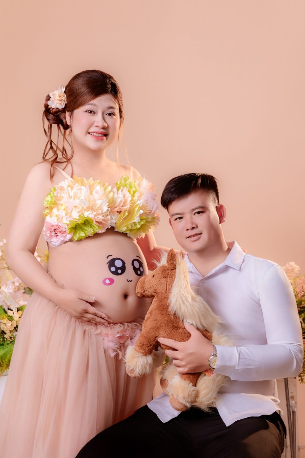 Chụp ảnh bầu cute với hình mặt cười mắt to má hồng trên bụng mẹ, mẹ mặc áo kết hoa và váy voan hồng đứng, chồng áo sơ mi trắng ngồi thấp hơn, ôm con ngựa bông